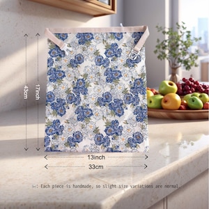 Op de afbeelding: Een rechthoekige opbergtas met bloemenpatroon, 43 cm hoog en 33 cm breed, met blauwe en witte bloemen op een cr&egrave;mekleurige achtergrond. De tas heeft beige bandjes en metalen gespen. De tekst luidt: "Elk stuk is handgemaakt, dus kleine maatvariaties zijn normaal."