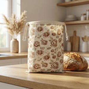 Pode incluir: Um saco de pão bege com um padrão de rosas castanhas, fecho de cordão e borda creme. O saco está sobre uma bancada de madeira ao lado de um pão.