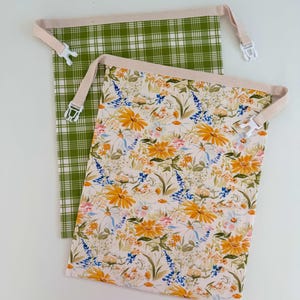 May include: Two drawstring bags. One bag has a green and white plaid pattern. The other bag has a floral pattern with yellow, white, and blue flowers on a cream background. Both bags have beige trim and white plastic buckles.