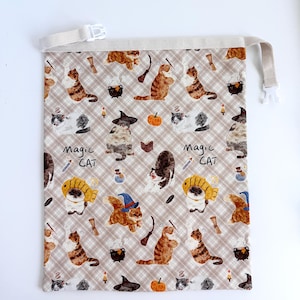 May include: Beige drawstring bag with a repeating pattern of cartoon cats, witch hats, cauldrons, and pumpkins. The words "Magic Cat" are printed on the bag. The bag has a beige strap and a white plastic clasp.