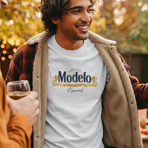May include: White t-shirt featuring the "Modelo Especial" logo in navy blue, gold, and red. The person is wearing a brown and tan jacket. The image is taken outdoors.