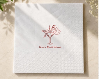 May include: A square white paper napkin featuring a red line drawing of a person wearing a veil and sunglasses, lounging inside a large martini glass while holding a drink. Below the illustration, the text reads, "Emma's Bridal Shower."