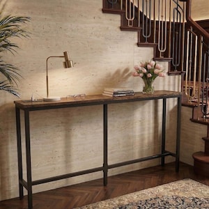 May include: A long, narrow console table with a dark brown wooden top and black metal frame. A gold-toned desk lamp, eyeglasses, and books sit on the table. A vase of pink tulips adds a touch of color. The table is 60 inches long.