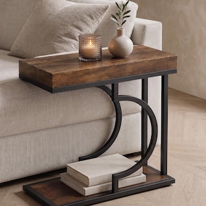 Op de afbeelding: Een moderne bijzettafel met een donkerbruin houten blad en een zwart metalen frame. De tafel heeft een onderste plank en een uniek gebogen ontwerp. Een kaars, een kleine vaas met groen en boeken staan op de tafel.