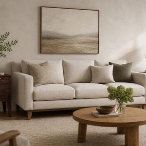 Puede incluir: Un sof&aacute; beige de tres cojines con dos mesitas de noche de madera oscura, una mesa de centro redonda de madera y un cuadro de paisaje enmarcado. El sof&aacute; tiene tapicer&iacute;a clara, mientras que las mesitas tienen tiradores dorados y follaje.
