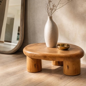 May include: A low, organic-shaped wooden coffee table with a smooth, rounded top and three thick, cylindrical legs. A tall, off-white textured ceramic vase containing thin, dried branches and a small, polished brass bowl sit on the table surface.