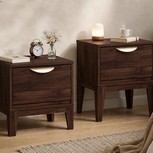 Puede incluir: Dos mesitas de noche de madera de color marr&oacute;n oscuro, cada una con un caj&oacute;n y un tirador curvo de tono dorado. Las mesitas se apoyan sobre cuatro patas c&oacute;nicas y est&aacute;n decoradas con objetos como un reloj, un jarr&oacute;n y una l&aacute;mpara.