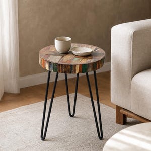 May include: A round side table with a tabletop constructed from multicolored reclaimed wood planks, supported by three slender black metal hairpin legs. A small off-white ceramic cup and a shallow matching dish sit on the table surface.