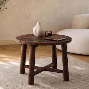 May include: A dark brown, round wooden side table with four sturdy legs and a cross-beam support. A white vase, a small candle, and a book rest on the table. The table sits on a neutral-colored rug.