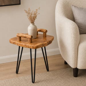 May include: A square wooden side table with a natural live edge, featuring two thick wooden handles on the top surface and four slender black metal hairpin legs. A light-colored ceramic vase containing dried flowers sits on the table.