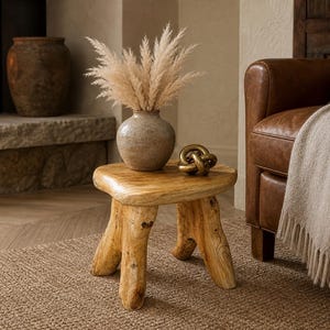 May include: A small, light-brown wooden stool with three thick, organic-shaped legs and a rounded triangular top. On the stool sits a round, speckled beige ceramic vase filled with fluffy, light-tan pampas grass and a small, gold-toned metal knot.