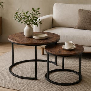 May include: A set of two round nesting coffee tables featuring dark brown wood-grain circular tops and black metal frames. The larger table holds a light-colored ceramic vase with green leaves and a small bowl, while the smaller table holds a cup.