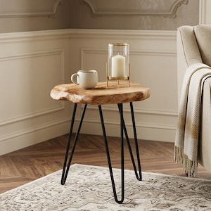 May include: A small, natural wood side table with black hairpin legs. A white mug and a candle in a glass holder sit on the table. The table is on a patterned rug in a room with a beige armchair.