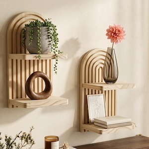 May include: Two wooden wall shelves with an arched design. One shelf holds a gray pot with a trailing plant and a wooden ring. The other shelf displays a vase with a pink flower and a framed picture. The shelves are mounted on a beige wall.
