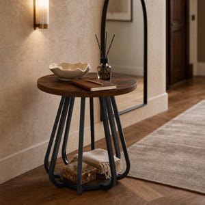 May include: A round side table with a dark wood grain top and a black metal frame featuring curved legs. On the table sits a cream-colored wavy bowl, a brown notebook, and a glass reed diffuser. A small box and a folded beige textile rest below.