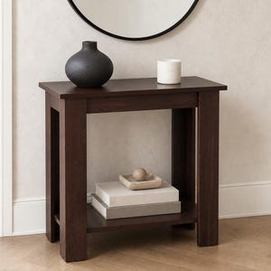 May include: A dark brown wooden console table with a rectangular top and a lower shelf. The top surface holds a matte black rounded vase and a white cylindrical candle. The lower shelf contains two stacked rectangular books and a small square dish.
