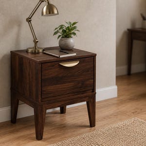 Puede incluir: Una mesita de noche de madera de color marr&oacute;n oscuro con un caj&oacute;n y un tirador curvo de tono dorado. La mesita se apoya sobre cuatro patas c&oacute;nicas y sostiene una l&aacute;mpara de escritorio color lat&oacute;n, una peque&ntilde;a planta verde y un cuaderno cerrado.