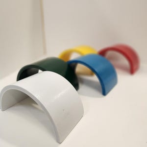 May include: A collection of six arched wooden toys in a rainbow of colors: white, green, yellow, blue, and red. The toys are arranged in a semi-circle, with the white arch in the foreground. The toys are likely for children.