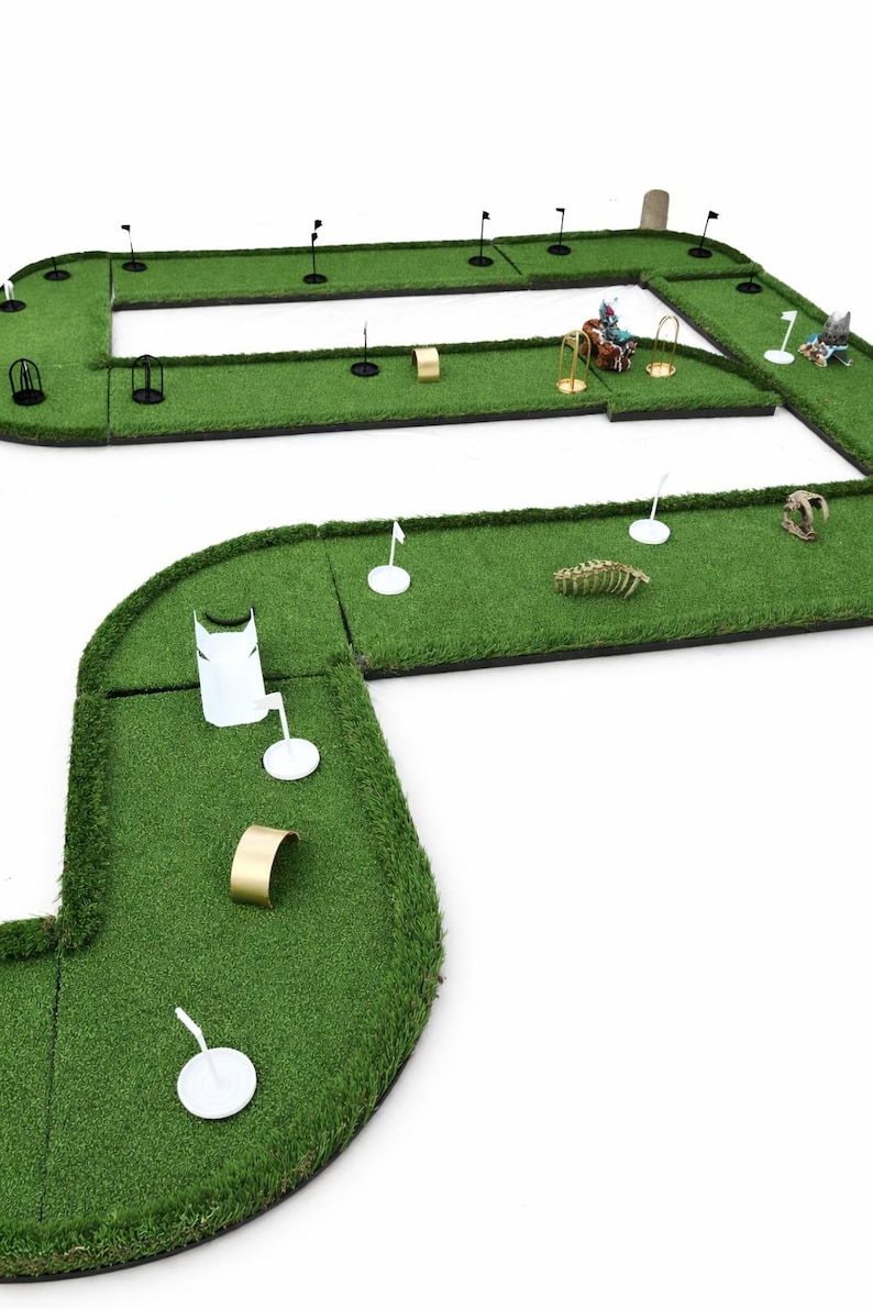 Puede incluir: Juego de minigolf en miniatura con c&eacute;sped verde, banderas negras y varios obst&aacute;culos. Incluye aros dorados, marcadores de tee blancos y elementos decorativos como un esqueleto de dinosaurio y figuritas.