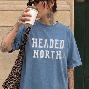 May include: A blue jean colored short-sleeve t-shirt with a crew neckline. The front of the shirt features the text "HEADED NORTH" printed in a distressed white font. The shirt has a relaxed fit and a solid, slightly faded blue appearance.