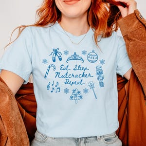 May include: Light blue t-shirt with a blue graphic design featuring ballet shoes, a tiara, ornaments, and the text "Eat. Sleep. Nutcracker. Repeat." The shirt is paired with light-wash jeans.