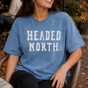 May include: A blue jean colored short-sleeve t-shirt featuring the white distressed text HEADED NORTH printed on the chest. The shirt has a crew neckline and a relaxed fit, with small decorative floral accents placed near the text lettering.