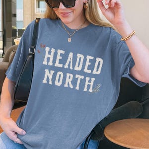May include: A blue-gray short-sleeve t-shirt featuring the text HEADED NORTH in a distressed white font across the chest. The shirt has a crew neckline and a relaxed fit, paired with a black shoulder bag, gold necklace, and gold bracelet.