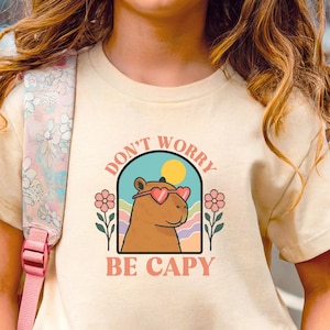May include: A cream-colored t-shirt featuring a cartoon capybara wearing heart-shaped sunglasses. The shirt has the text "DON'T WORRY BE CAPY" in a retro design with flowers and a sun. The word "NATURAL" is printed at the bottom.