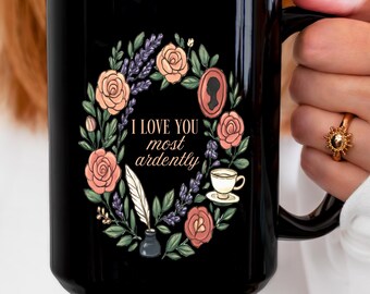 May include: Black ceramic mug featuring a floral wreath design with roses, lavender, and the text "I love you most ardently." The design includes a cameo, teacup, and quill. The mug is held by a person.