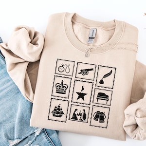 Peut inclure: Sweat-shirt couleur sable avec un motif de timbres noirs représentant une horloge, un canon, une plume, une couronne, une étoile, un piano, un navire, un couple et des chopes de bière. Le mot "SAND" est imprimé en bas à droite. Un collier argenté est drapé sur le sweat-shirt.