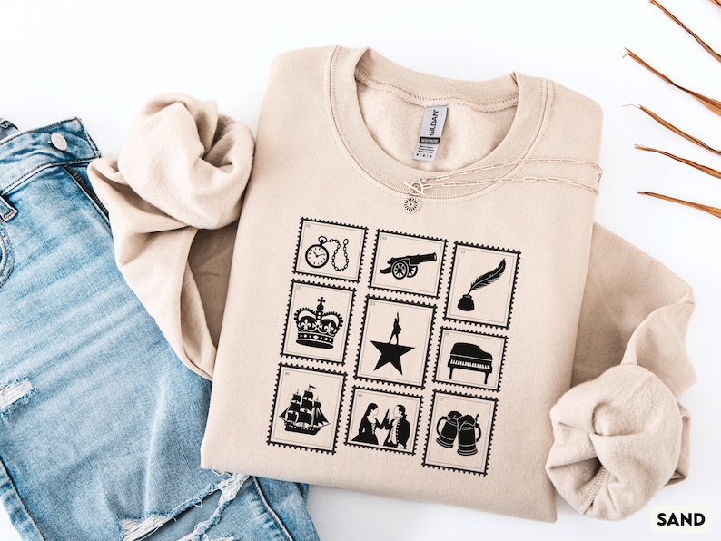 Puede incluir: Sudadera de color arena con un dise&ntilde;o de sellos negros que presenta un reloj, un ca&ntilde;&oacute;n, una pluma, una corona, una estrella, un piano, un barco, una pareja y jarras de cerveza. La palabra "SAND" est&aacute; impresa en la esquina inferior derecha. Un collar plateado est&aacute; drapeado sobre la sudadera.