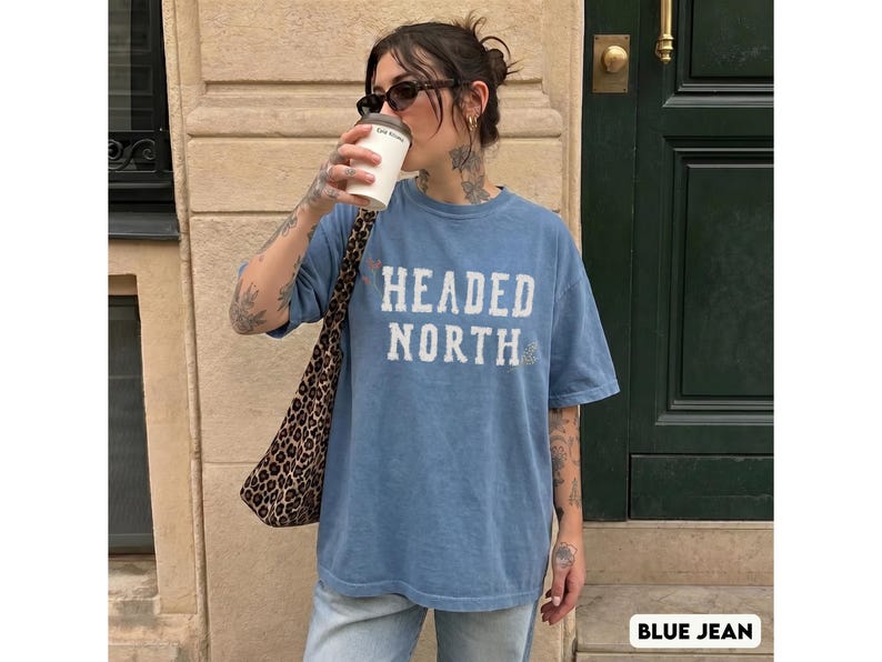 May include: A blue jean colored short-sleeve t-shirt with a crew neckline. The front of the shirt features the text "HEADED NORTH" printed in a distressed white font. The shirt has a relaxed fit and a solid, slightly faded blue appearance.