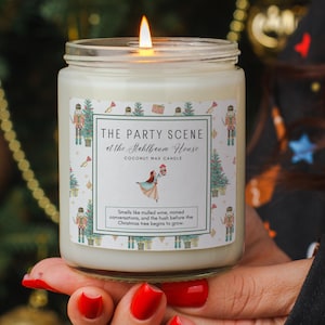 May include: A lit coconut wax candle in a glass jar. The label reads "THE PARTY SCENE at the Stahlbaum House." The label also includes a graphic of a ballerina and a Christmas tree. The candle is held in a person's hand.