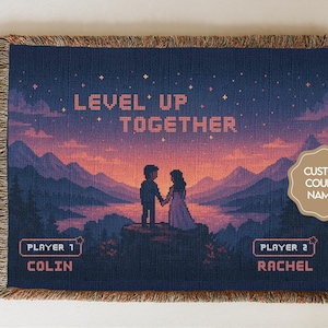 May include: A woven blanket with a pixelated design showing a couple holding hands against a mountain backdrop. The text "LEVEL UP TOGETHER" is above them. Below, it says "PLAYER 1 COLIN" and "PLAYER 2 RACHEL".