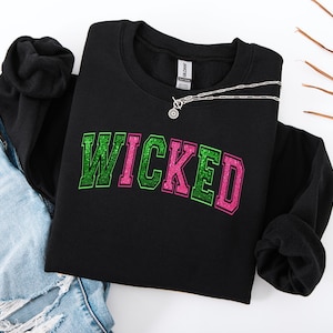 Peut inclure: Sweat-shirt noir avec le mot "WICKED" en lettres pailletées vertes et roses. Un collier argenté avec un pendentif circulaire est posé sur le sweat-shirt. Le sweat-shirt est associé à un jean bleu.