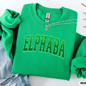 May include: A green sweatshirt with the name "ELPHABA" in a glittery, collegiate-style font. The sweatshirt is a vibrant Irish green colour. A silver necklace with a circular pendant is draped across the top. Blue jeans are visible in the background.