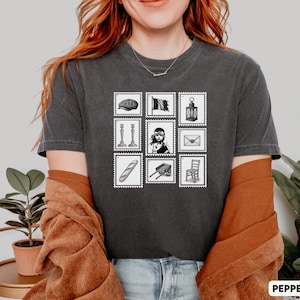 Puede incluir: Camiseta gris oscuro con una cuadrícula de nueve ilustraciones estilo sello postal. Los sellos representan una boina, una bandera, una linterna, candelabros, un retrato, un sobre, una baguette, un carro y una silla. La palabra "PEPPER" está en la parte inferior.