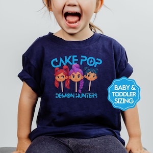 May include: Navy blue t-shirt with the text "CAKE POP DEMON HUNTERS" in blue. The graphic features three cartoon cake pops with different hair colors. A blue badge indicates baby and toddler sizing.