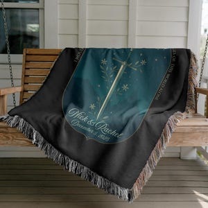 Op de afbeelding: Zwarte geweven sprei met een blauwgroen en goudkleurig ontwerp met een zwaard, bloemaccenten en de namen "Nick & Rachel" met een datum. De deken heeft een gefranjerde rand en is over een houten schommelbank gedrapeerd.