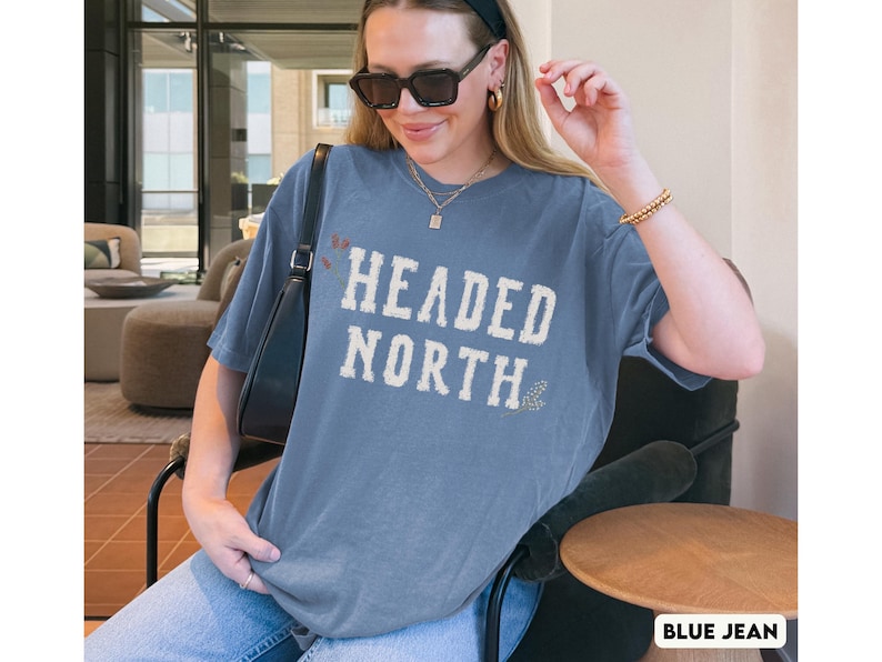 May include: A blue-gray short-sleeve t-shirt featuring the text HEADED NORTH in a distressed white font across the chest. The shirt has a crew neckline and a relaxed fit, paired with a black shoulder bag, gold necklace, and gold bracelet.