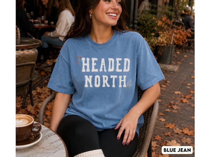 May include: A blue jean colored short-sleeve t-shirt featuring the white distressed text HEADED NORTH printed on the chest. The shirt has a crew neckline and a relaxed fit, with small decorative floral accents placed near the text lettering.