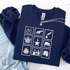 Puede incluir: Sudadera azul marino con un dise&ntilde;o de sellos postales blancos. Los sellos representan un reloj, un ca&ntilde;&oacute;n, una pluma, una corona, una estrella, un piano, un barco, figuras y jarras de cerveza. Un collar plateado con un colgante est&aacute; sobre la sudadera.