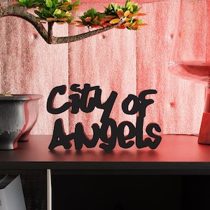 Op de afbeelding: Een zwart houten bord met de tekst "City of Angels" in cursiefschrift staat op een donkere plank. Links een bonsai-boom in een donkere pot. Rechts een rood getinte lamp. Boeken zijn eronder zichtbaar.