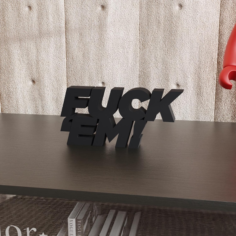 May include: A black, three-dimensional sculpture spells out "FUCK EM!" in bold, sans-serif letters. The sculpture is placed on a dark brown surface, with a textured, light-colored wall in the background. The letters are angled and stacked for a dynamic look.