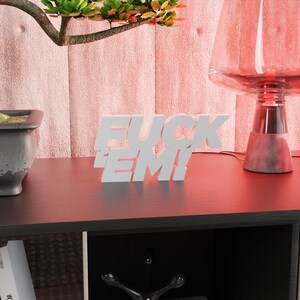 May include: A white, illuminated sign with the text "FUCK EM!" is displayed on a dark surface. The sign is in a bold, sans-serif font. A bonsai tree in a gray pot and a red-tinted lamp are also visible.