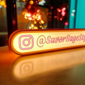 May include: A rectangular, illuminated sign with a pink Instagram logo and the text "@SavorSageStyle". The sign has a warm, orange glow and is set against a blurred background with bokeh lights. The sign is likely used for social media promotion.