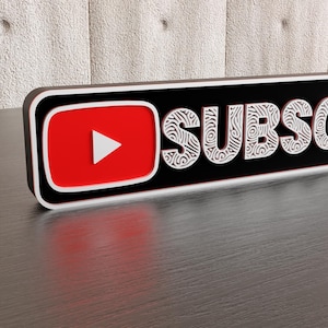 Könnte beinhalten: Ein rechteckiges Schild mit dem Wort "SUBSCRIBE" in weißen, verzierten Buchstaben. Die linke Seite zeigt das YouTube-Logo in Rot mit einer weißen Wiedergabetaste. Das Schild hat einen schwarzen Hintergrund und einen weißen und schwarzen Rand.