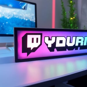 Könnte beinhalten: Ein rechteckiges, beleuchtetes Schild mit dem Twitch-Logo und dem Wort "YOURNAME" in Weiß, vor einem rosa-violetten Farbverlauf. Das Schild befindet sich in einem schwarzen Rahmen und steht auf einer weißen Oberfläche.