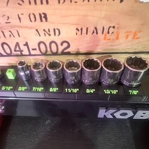 May include: A set of chrome sockets of various sizes, ranging from 1/4" to 7/8", displayed on a black holder. The sizes are labeled in green text. The background includes a wooden surface with faded text.