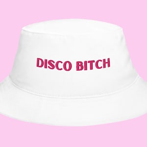 May include: White bucket hat with the text "DISCO BITCH" in a bold, pink font. The hat is made of a soft material and has a classic bucket hat shape. The background is a light pink color.